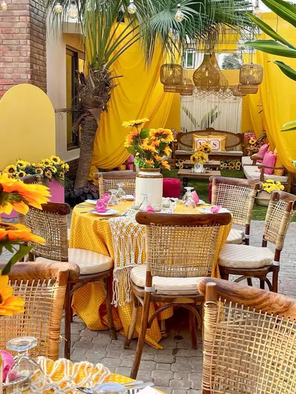 A dining setup for an intimate Haldi lunch. The tables are styled with polka-dot yellow tablecloths, macrame runners, and cane chairs to perfectly match the bohemian, sunny theme.