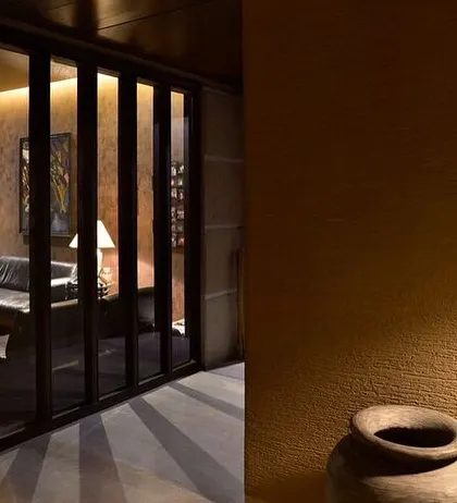 A glimpse into a living space through a screen of vertical wooden slats. The warm, low lighting creates an intimate and moody atmosphere, hinting at a comfortable room beyond.