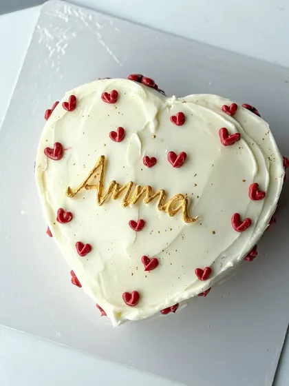 A simple and heartfelt cake for Amma. This heart-shaped cake is covered in white buttercream and decorated with tiny red hearts and a beautiful gold "Amma" script.