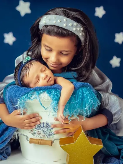 A big sister lovingly holds her sleeping newborn sibling against a starry blue backdrop. A magical and heartwarming newborn and sibling portrait.