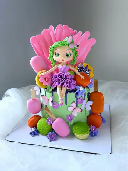 A vibrant and colorful cake that's full of life. This design features a green drip, macarons, and a cute fairy doll, suggesting a fun and playful theme.