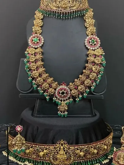 This bridal set combines green beads with traditional kemp stones for a vibrant look. It includes a choker, a long haaram, and a matching vaddanam, perfect for coordinating with green or red sarees.