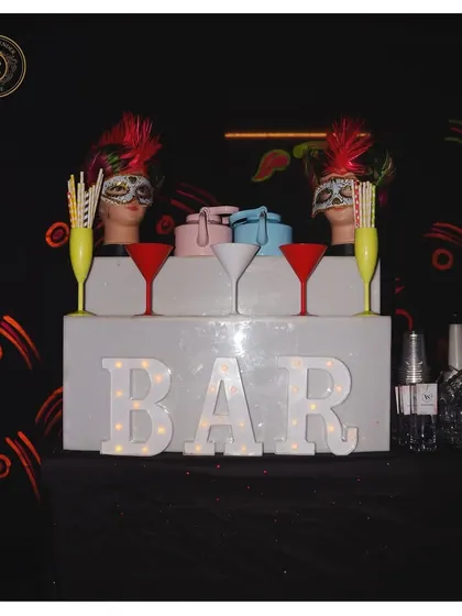 A minimalist bar display with a touch of carnival flair. The masked mannequins and bold martini glasses create a striking look against the simple, illuminated bar sign.