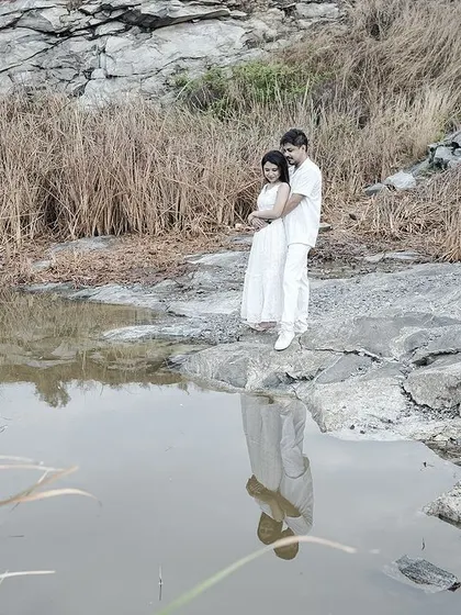 A wide shot of a couple embracing by a quarry lake, their reflection in the water, set against a rustic, natural backdrop.