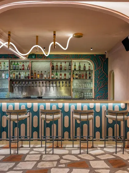 The bar at Hangover features a playful design with a custom-patterned counter, teal wallpaper, and a unique, wave-like neon light fixture. The crazy-paving terrazzo floor adds another layer of texture and color to this eclectic space.