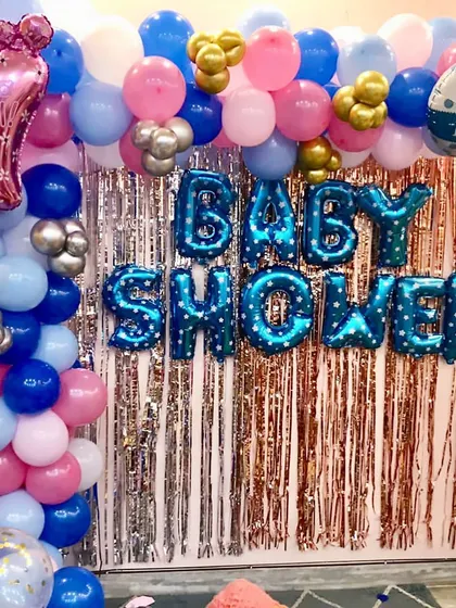 A simple home setup for a baby shower. A silver foil curtain is framed by a pink and blue balloon arch, with "BABY SHOWER" foil letters.