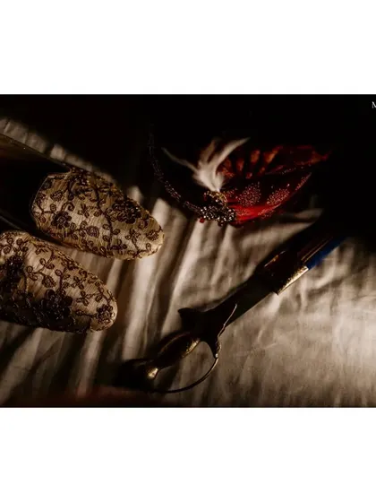 A moody, artistic shot of the groom's wedding accessories. The traditional sword, turban feather, and embroidered shoes are laid out, capturing the regal details in a dramatic play of light and shadow.