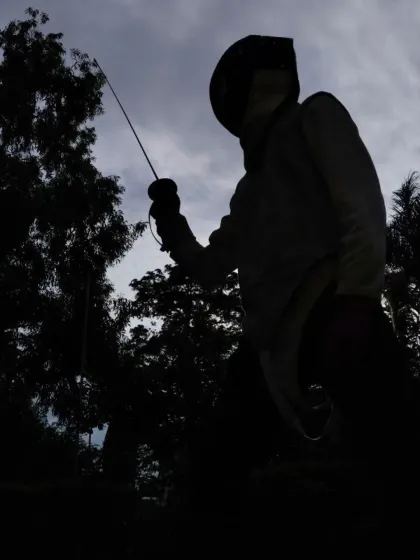 A powerful, low angle silhouette of a fencer in a ready stance. The dramatic lighting highlights the warrior-like posture and readiness for combat.