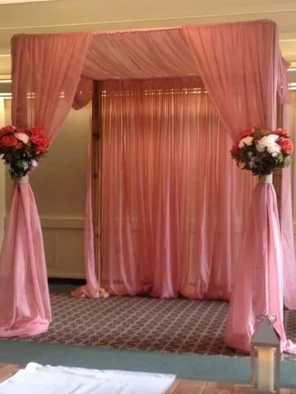 A simple and elegant mandap design suitable for an indoor wedding or an intimate ceremony. The structure is draped with sheer pink fabric and accented with two beautiful floral bouquets, creating a sacred space that is both modern and graceful.