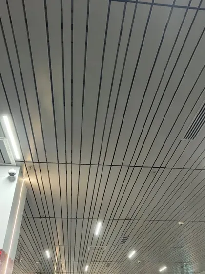 A modern, linear metal ceiling in a commercial space. The sleek white panels with black grooves integrate lighting and air conditioning vents for a clean, minimalist finish.
