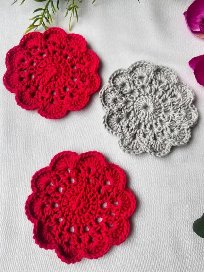 A set of two red and one grey mandala coasters, laid out to show the beautiful floral-inspired pattern. Each one is handmade with 100% cotton yarn.