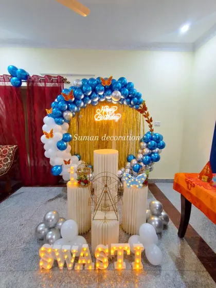 A butterfly-themed birthday setup with a blue, white, and silver balloon ring against a gold tinsel curtain. The name 'Swasti' is displayed in marquee letters.
