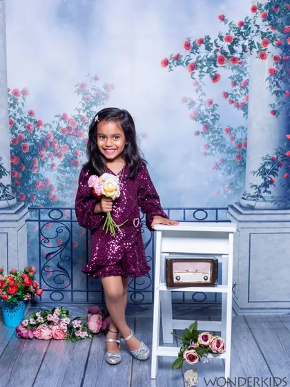 A smiling girl in a sparkly purple dress holds a bouquet of flowers in a romantic, floral-themed setting. The backdrop of pillars and roses adds a touch of elegance.