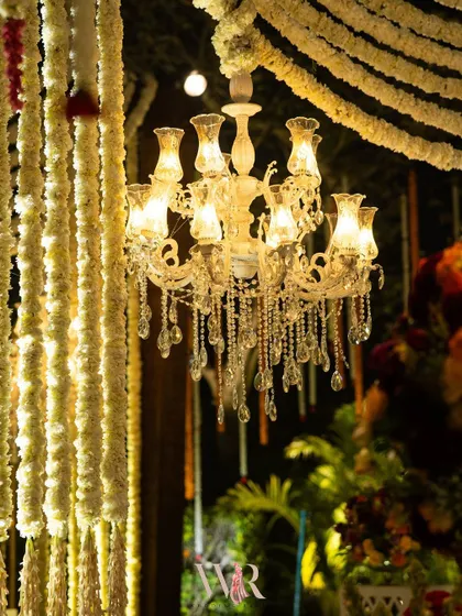 A close-up of a crystal chandelier against a curtain of traditional white floral garlands. This juxtaposition of classic elegance and cultural heritage is a signature of our design philosophy.