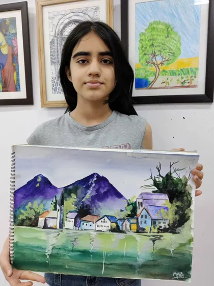 A young artist shows her completed watercolor landscape, featuring purple mountains and a reflective lake. This piece highlights her progress in creating atmospheric depth.