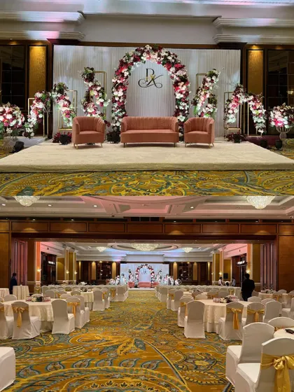 A full view of a ballroom setup, showing how the stage design with its floral arch and monogram integrates with the overall guest seating and venue architecture.