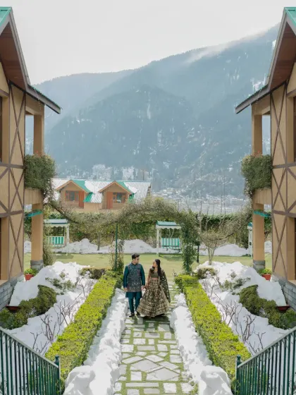 A stunning wide shot of the couple walking in a manicured garden with snow and mountains in the background, capturing the magic of their Manali Mehendi.