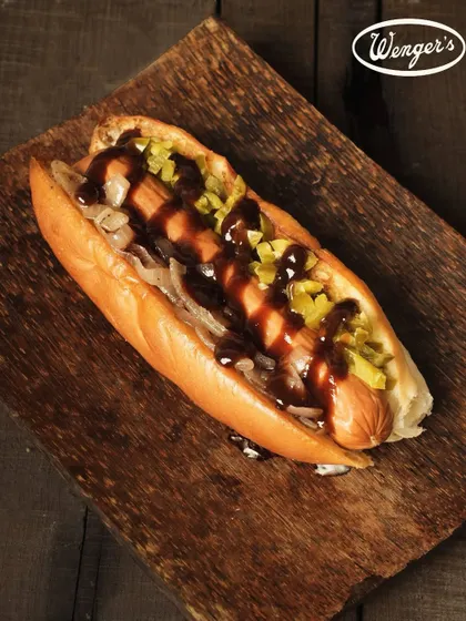 Our take on the classic Hot Dog. This image shows a juicy sausage nestled in a soft, fresh bun from our bakery, topped with onions, relish, and a generous drizzle of sauce.