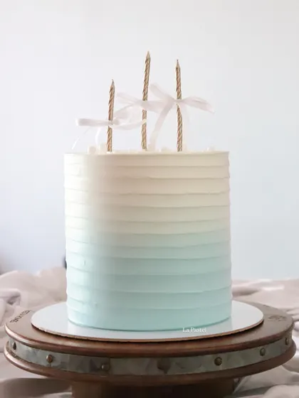 A tall and elegant ombre cake, transitioning from a soft blue to white. The simple design is finished with three candles tied with delicate white ribbons.