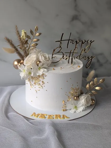 A minimalist and elegant white and gold birthday cake. The clean white finish is decorated with a sprinkle of gold leaf, white roses, and delicate dried florals for a touch of class.