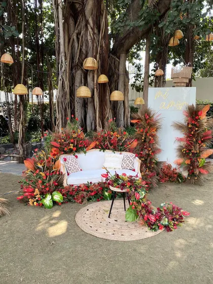 A cozy and picturesque photo booth nestled under a large banyan tree. A comfortable sofa is surrounded by an explosion of autumnal florals, while hanging wicker lamps add a touch of rustic charm.