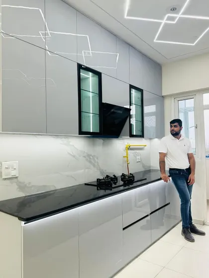 Posing in a newly completed grey modular kitchen. The design features a marble-finish backsplash, a black chimney, and glass-fronted overhead cabinets, combining functionality with high-end aesthetics.