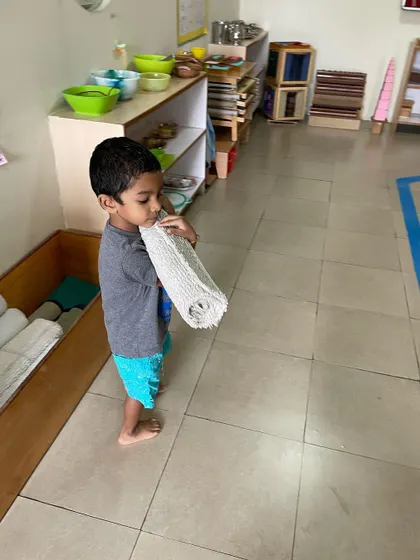 Carrying the rolled mat back to its storage place is the final step in this work cycle, teaching completion and care for the classroom materials.