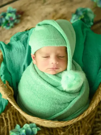 A perfect little bundle. This classic newborn pose shows a baby swaddled in a soft green wrap, nestled peacefully in a rustic basket. The pom-pom hat adds a touch of playful charm.