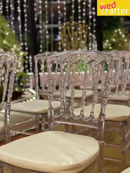 A close-up of the elegant transparent chairs. Their intricate design adds a touch of sophistication, especially when set against a curtain of warm, twinkling lights.