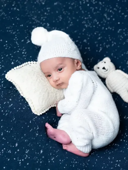 A newborn boy in a white knit romper and hat, looking thoughtful while lying on a starry night background.