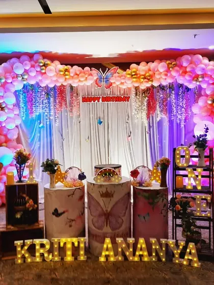 A stunning butterfly-themed setup for Kriti Ananya. The decor features a dramatic balloon arch with hanging floral strings and large butterfly props against a white curtain backdrop.