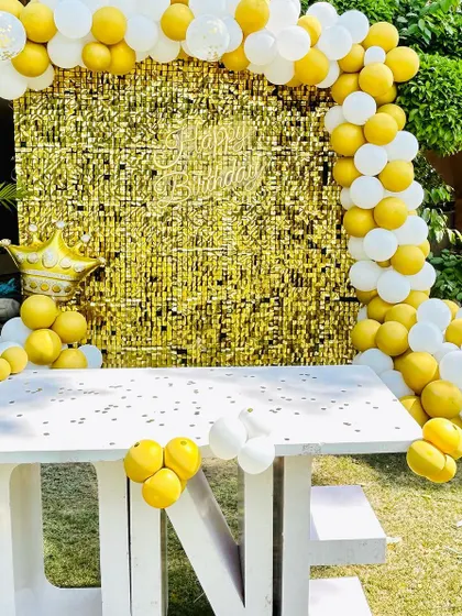 A simple and sweet outdoor birthday setup. A white "ONE" table is placed in front of a gold shimmer curtain, framed by a yellow and white balloon arch.