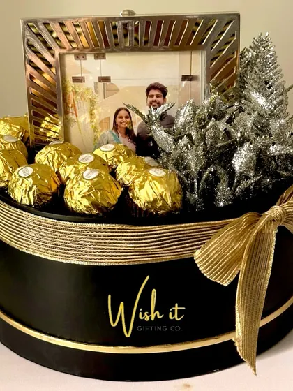 A sophisticated wedding hamper in a round black and gold box. It features a photo frame with the couple's picture and is surrounded by Ferrero Rocher chocolates, creating a personalized and luxurious gift.