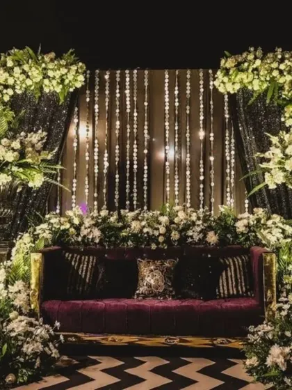 An opulent Sangeet seating area with a plush purple sofa, a backdrop of shimmering crystals, and large arrangements of white flowers.