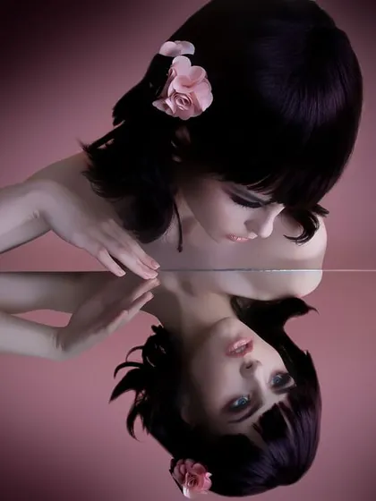 A creative beauty portrait showing a model with a sleek black bob and a pink flower in her hair, gazing at her own reflection in a mirror. This image showcases my technical skill with lighting and composition for artistic and conceptual photoshoots.