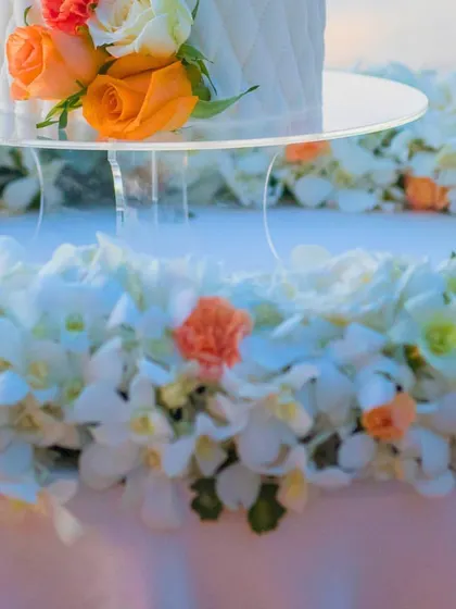 A close-up of the wedding cake, resting on a clear stand surrounded by a bed of white and orange flowers. It's these small, elegant details that elevate the entire look.