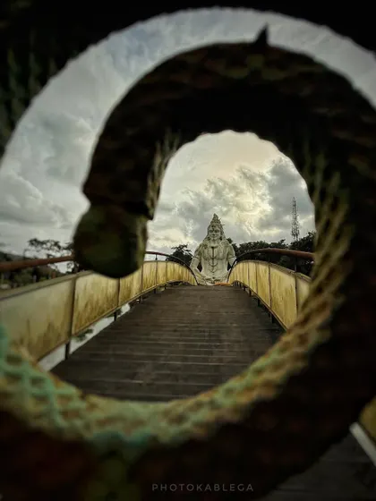 A creative shot of the Barfeshwar Mahadev statue in Virar, framed through the curve of a serpent sculpture on the bridge.