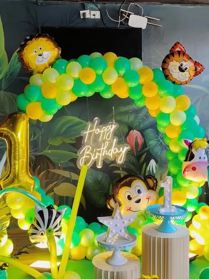 A jungle themed first birthday setup with a neon 'Happy Birthday' sign, animal foil balloons, and themed cake plinths against a leafy backdrop.