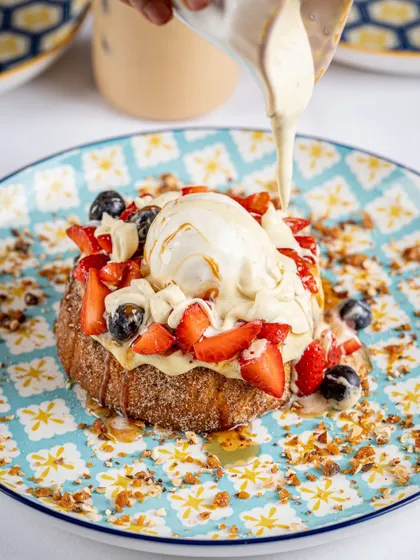 A close-up shot of cream being poured over a berry-topped French toast. The motion and glistening syrup make this an incredibly appetizing shot, perfect for a brunch menu.