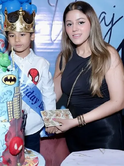 The birthday boy, dressed as a superhero with a crown, stands proudly by his multi-tiered superhero-themed cake.