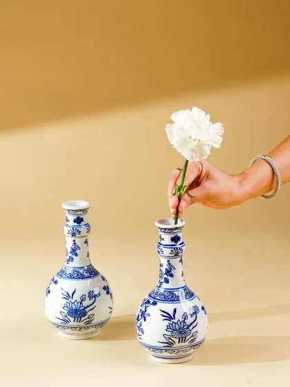 A hand places a white carnation into a blue and white ceramic vase. This simple, elegant action shot adds a human element and a touch of grace to the product photography.