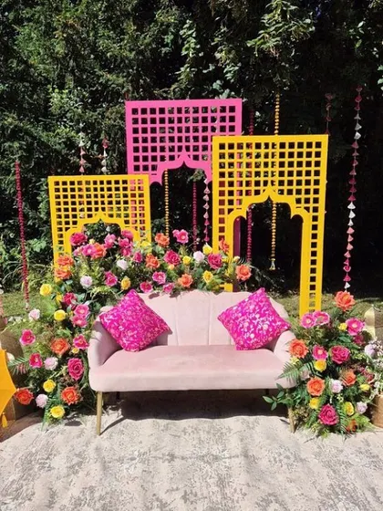 A vibrant outdoor Mehendi setup with pink and yellow lattice panels. The lush floral arrangements and colorful umbrellas make this a beautiful spot for photos.