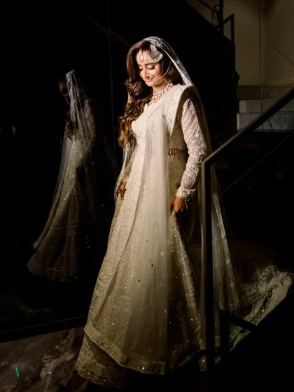A full-length portrait of a bride in a magnificent white lehenga, her reflection captured in the glossy wall beside her. The dramatic lighting highlights the intricate details of her outfit.