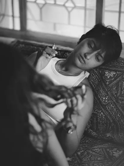 A relaxed, candid black and white photo of a woman leaning back on a patterned rug, a cigarette in hand. The over-the-shoulder perspective adds to the intimate, unposed feel of the moment.