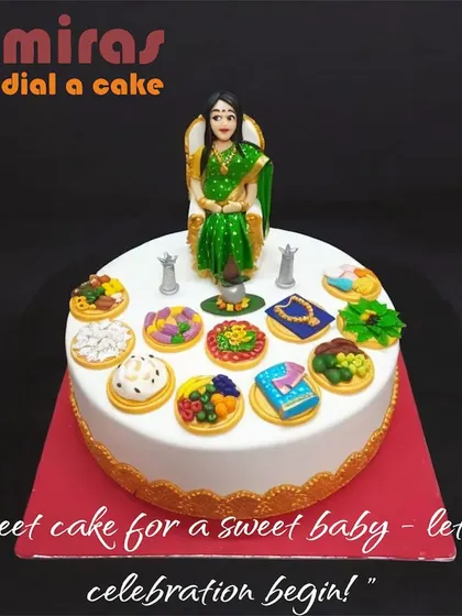 An elegant and traditional baby shower cake. This design features a figurine of a mother-to-be in a beautiful saree, surrounded by symbolic items for the ceremony.