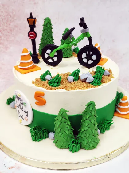 We set the scene for an outdoor adventure with this cycle theme cake. A green bicycle is parked on a dirt path surrounded by pine trees and traffic cones, creating a fun and detailed landscape for a young cyclist's birthday.