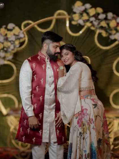 A happy portrait of a couple during their engagement or Sangeet. The bride leans affectionately on the groom's shoulder, both smiling in front of a beautifully decorated floral backdrop.