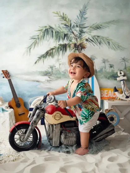 A happy toddler pretends to ride a miniature motorcycle in a fun, beach-themed studio session.