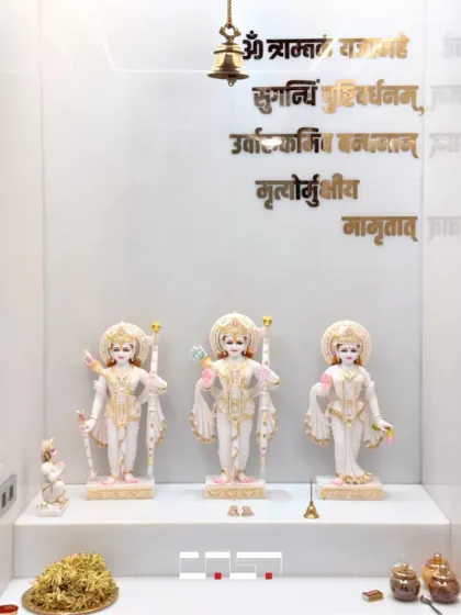 A close-up of the beautiful marble idols of Ram, Sita, and Lakshman, set against a pristine white backdrop with a hanging brass bell, creating a pure and peaceful focal point.