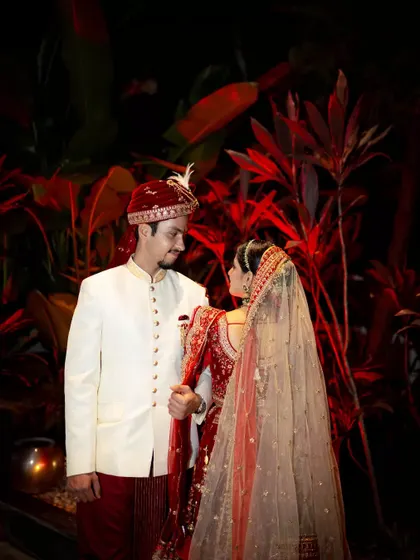 A classic wedding portrait of a couple in their red and white traditional attire, set against a dramatic, dark background.